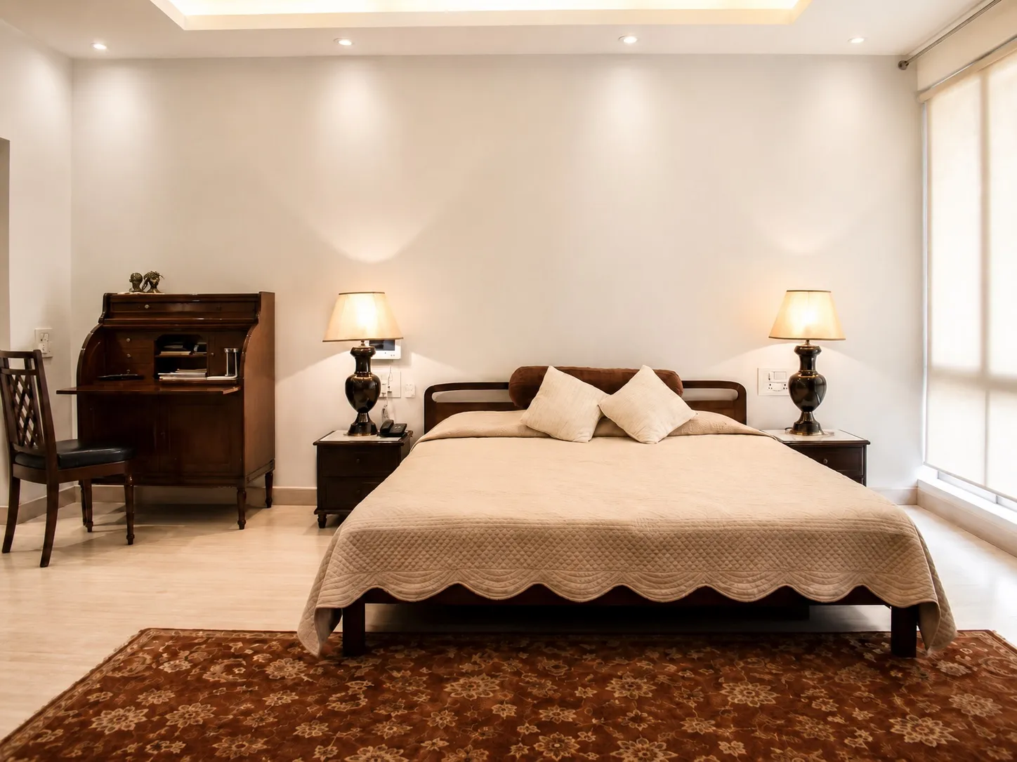 Master bedroom with an antique walnut roll-top writing desk, twin black urn table lamps, a beige quilted bed, and a deep-red floral rug