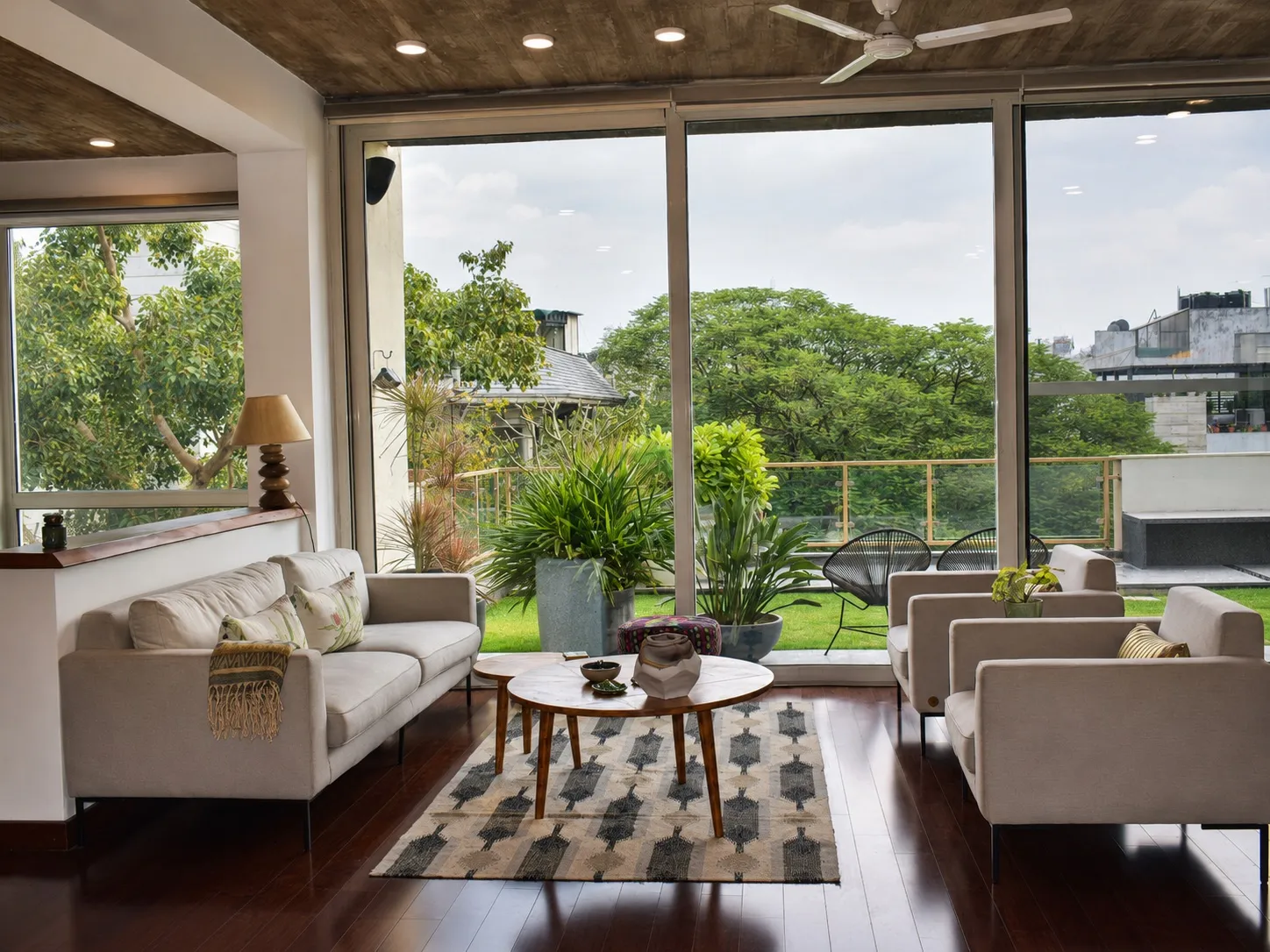 Living room with floor-to-ceiling glass opening to a green terrace, a pale-grey sofa and armchairs, a round marble-and-wood coffee table, and a patterned dhurrie on dark hardwood