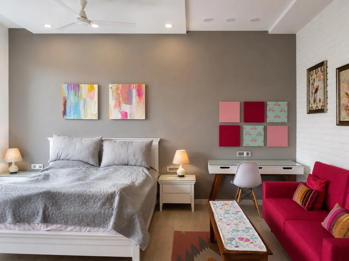 Child's bedroom with grey walls, colourful abstract paintings over a white bed, a magenta sofa, a study desk, flamingo-print wall panels, and a white-brick accent wall