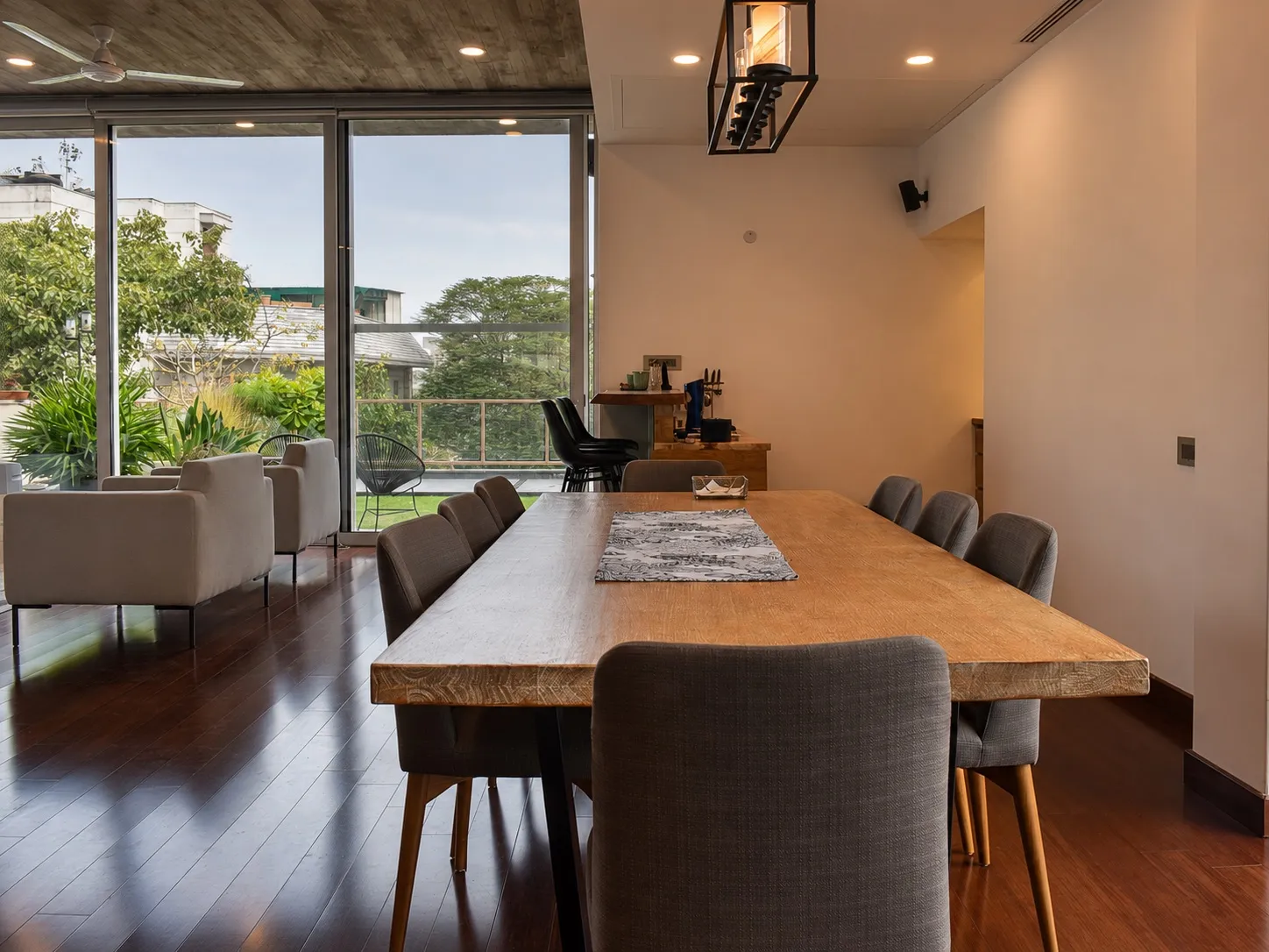 Dining area with a live-edge wood table and grey upholstered chairs, a black caged lantern pendant, and floor-to-ceiling sliding glass opening to a planted terrace
