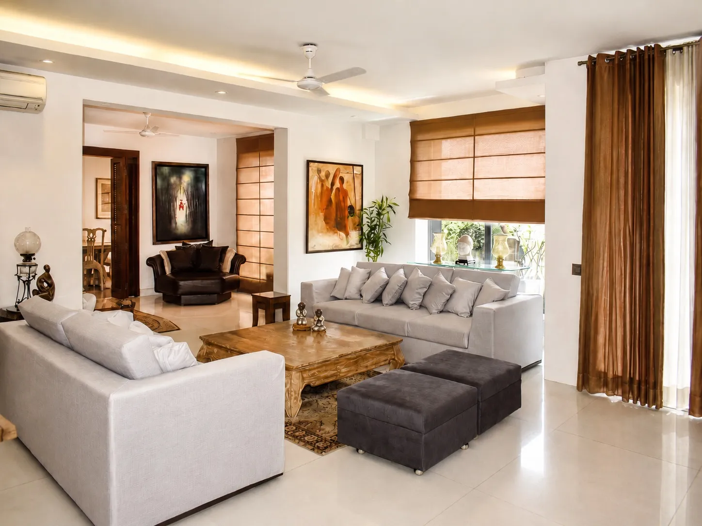 Living room with two pale-grey sofas, a hand-carved gold-wood coffee table, charcoal suede ottomans, brown drapes, and contemporary Indian paintings