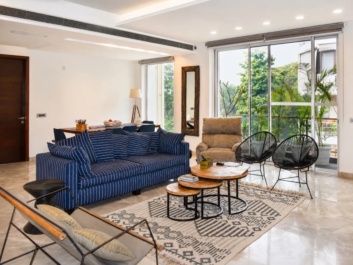 Living room with a navy-and-white striped sofa, round wooden nesting coffee tables, black Acapulco chairs, a patterned rug, and floor-to-ceiling glass opening to a green balcony
