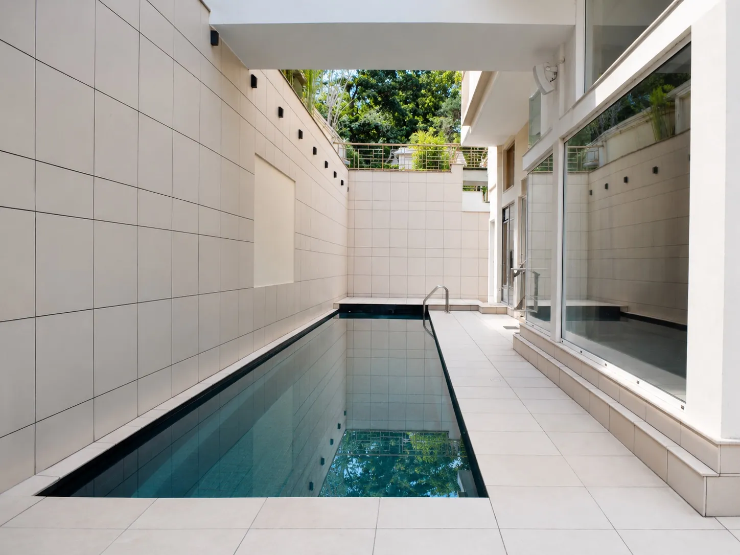 Narrow outdoor lap pool set between cream stone-tiled walls, running alongside the bungalow's glazed windows