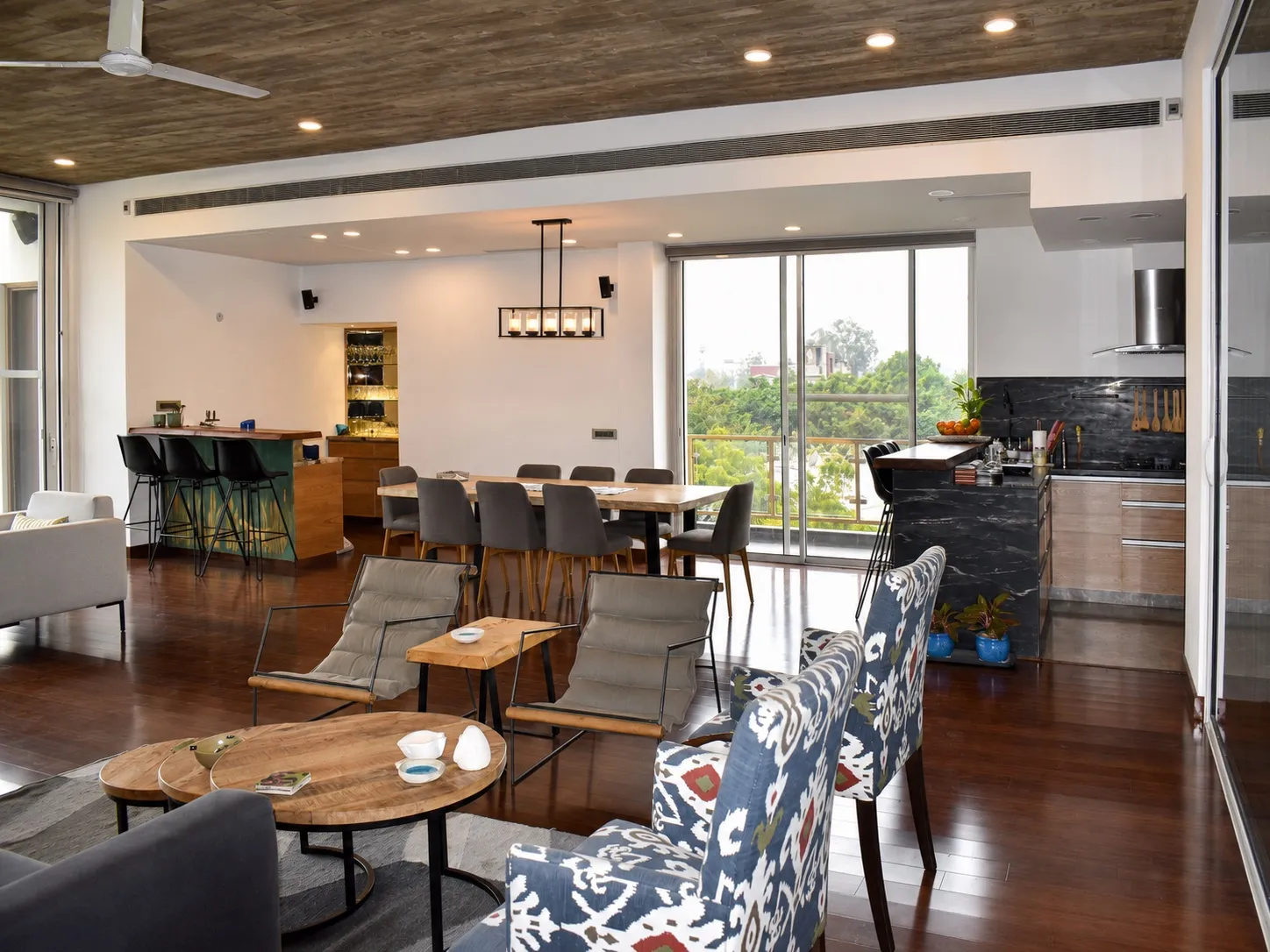 Open-plan living, dining and kitchen with a live-edge dining table under a linear pendant, a home bar with black stools, and a black-marble kitchen island with a chimney