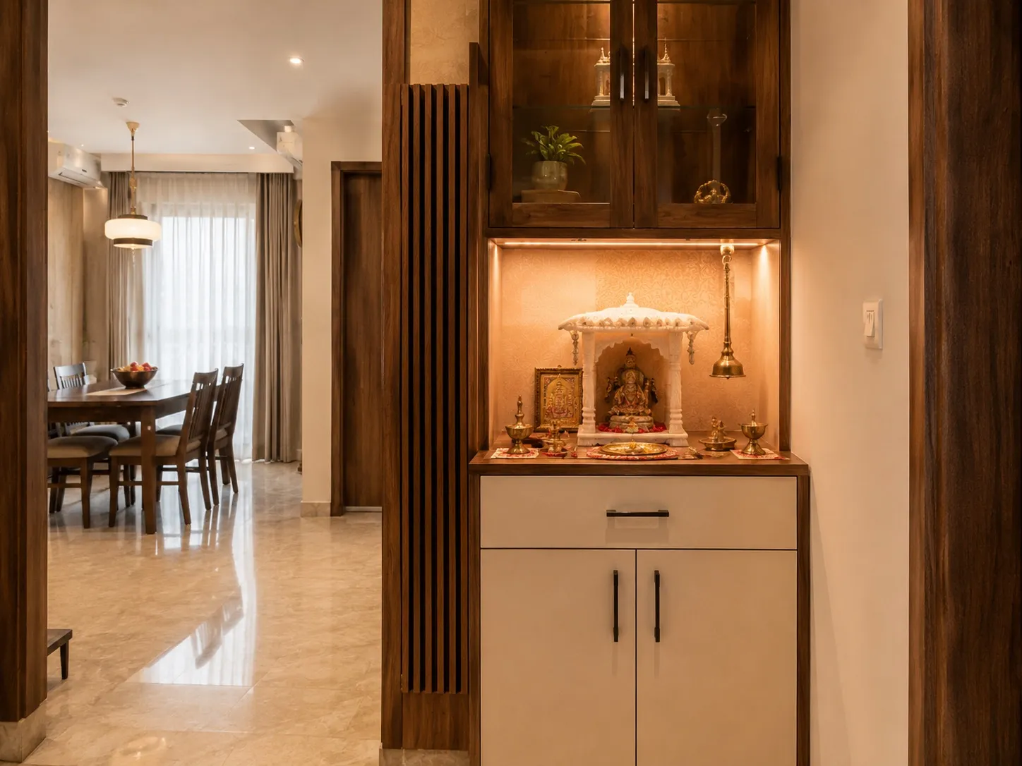Pooja unit with a carved white-marble temple in a backlit niche, brass lamps and a bell, a glass-front display cabinet above and a white-and-walnut base cabinet, beside a fluted-wood screen with the dining room beyond