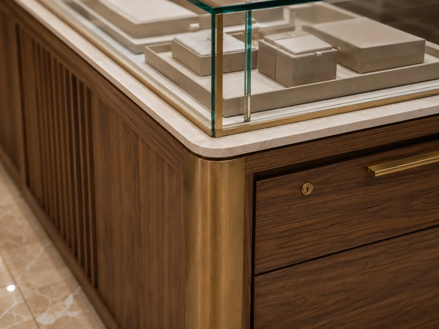 Close-up of a display counter corner in reeded walnut with a rounded brushed-brass trim, a cream-marble top, a brass-framed glass vitrine of beige trays, and a lockable drawer with a brass handle