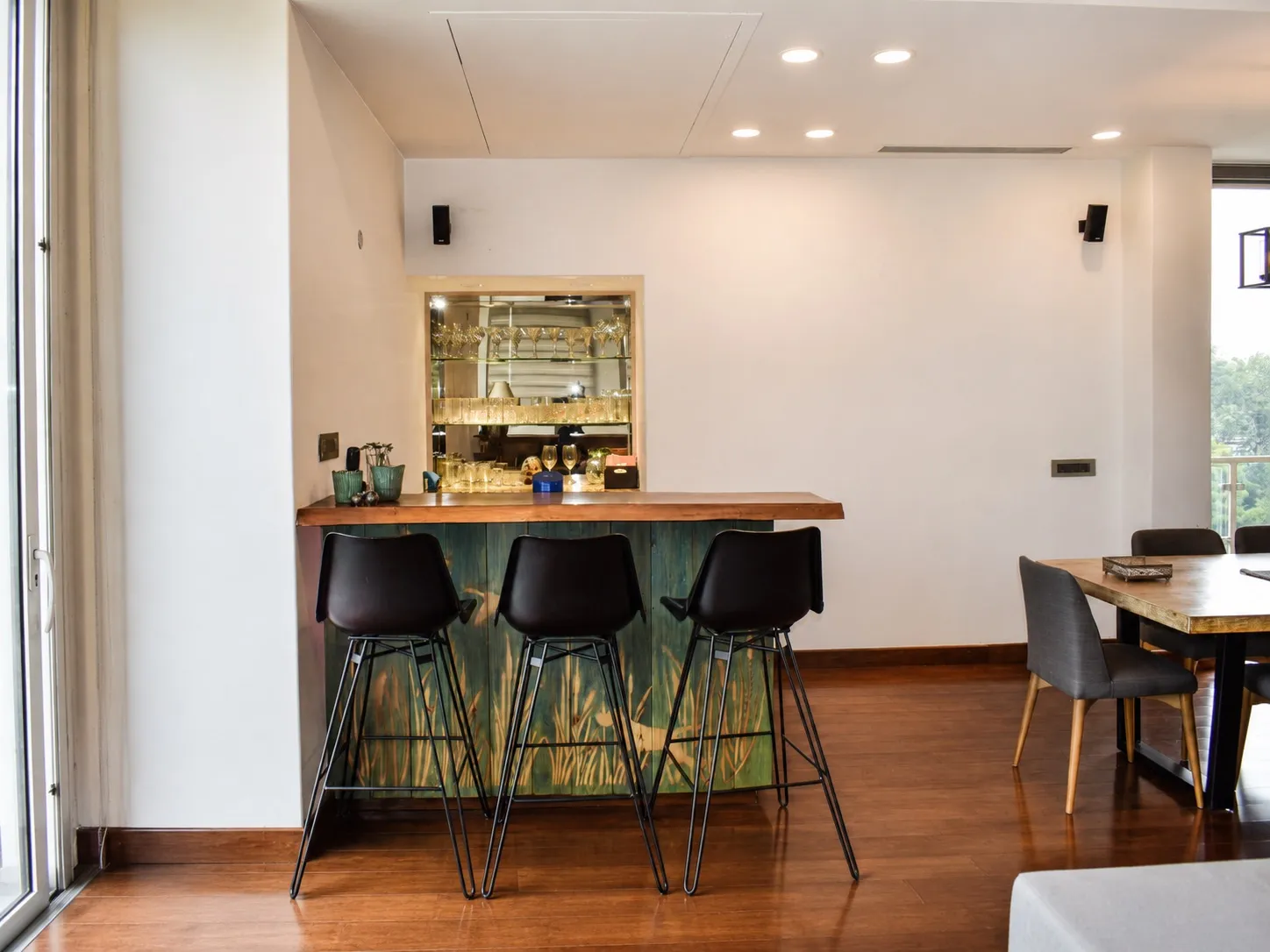 Home bar with a live-edge wood counter on a green painted base, three black leather bar stools, and a mirrored backlit niche displaying glassware