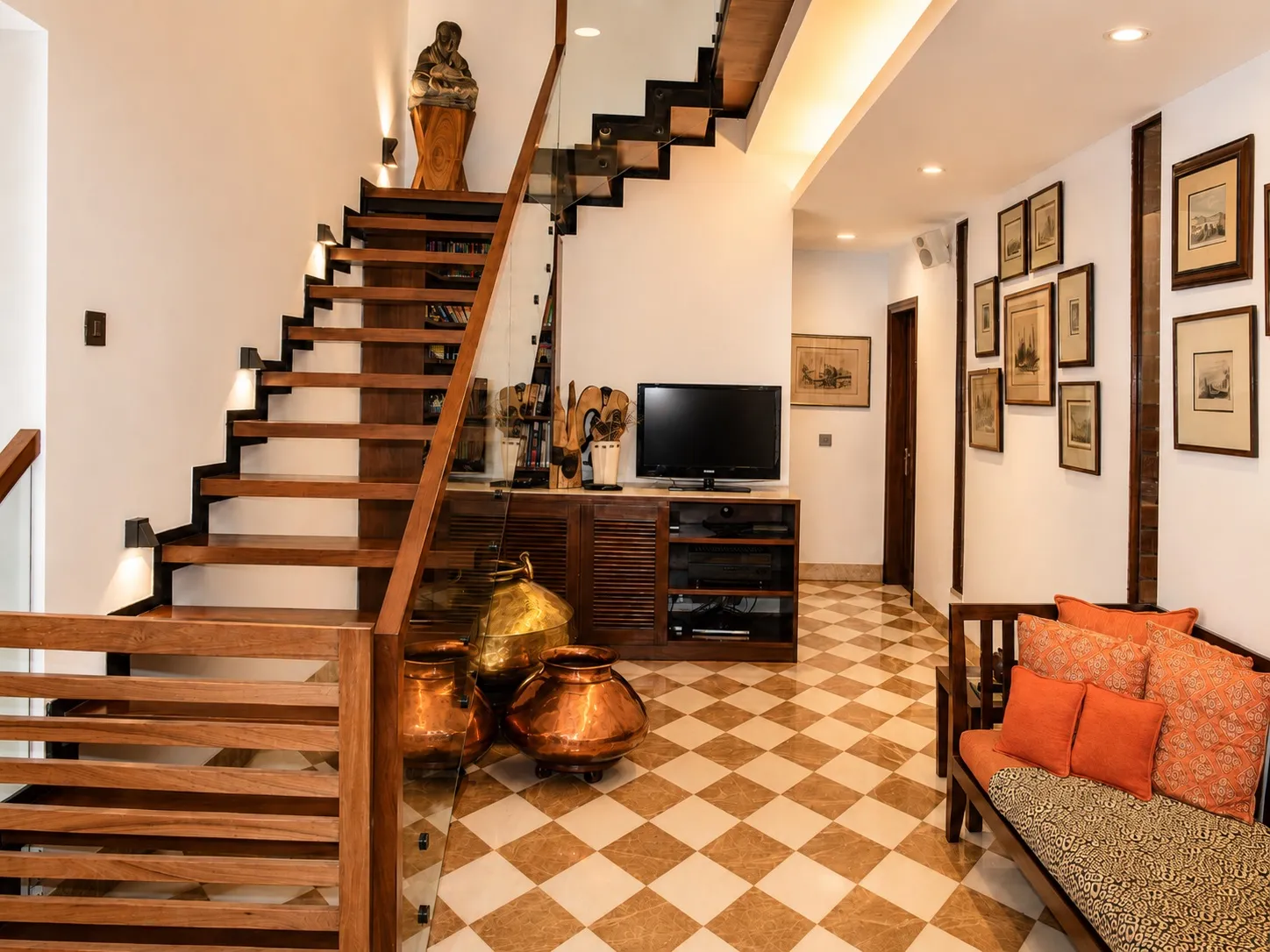 Walnut and glass staircase beside a TV lounge with brass and copper matka pots, a framed gallery wall, and an orange-cushioned daybed on a checkered marble floor