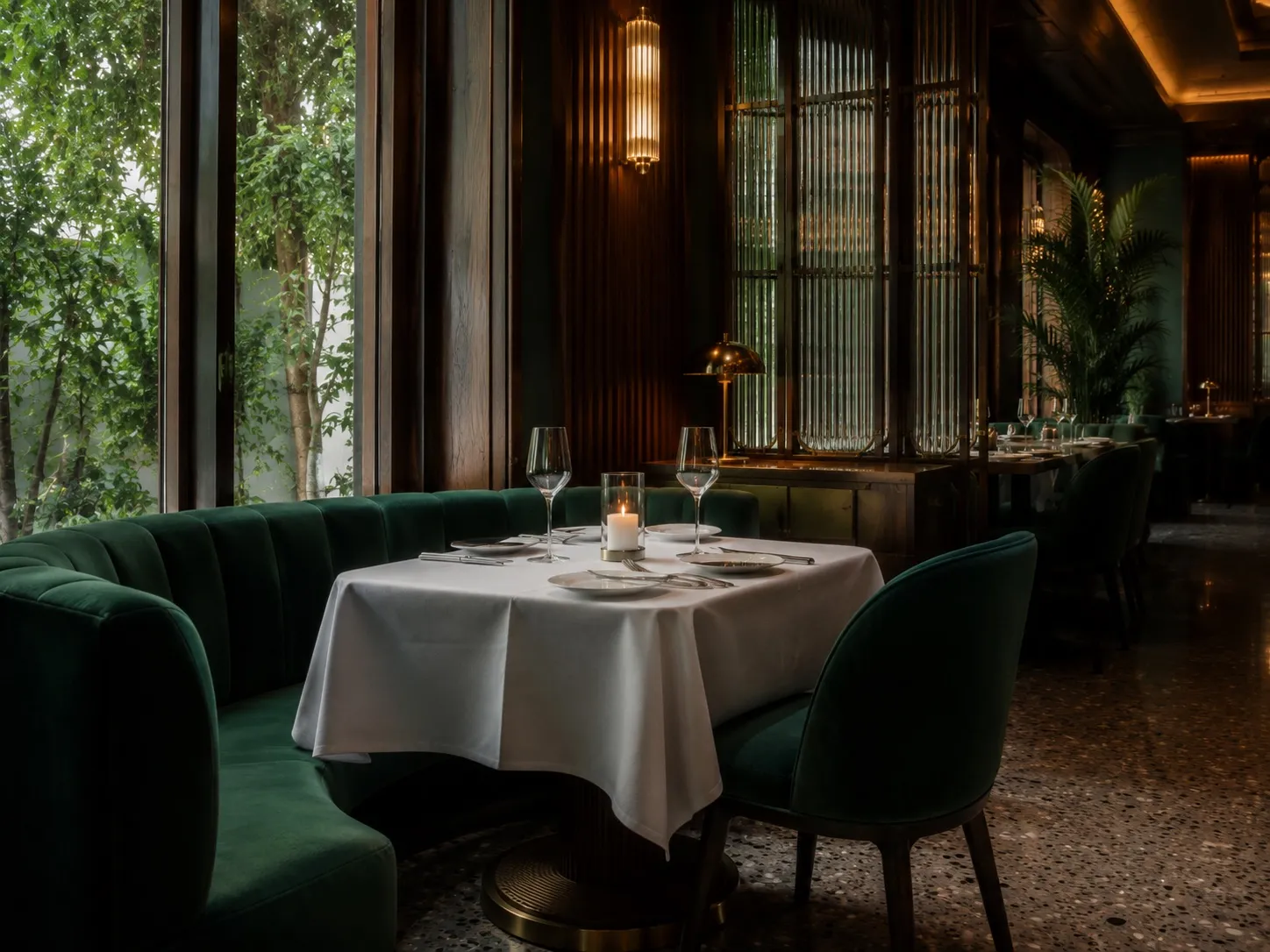 Window-side dining booth with a curved emerald velvet banquette and chair, a white-linen table set with wine glasses and a candle, fluted-glass screens, and greenery through floor-to-ceiling windows