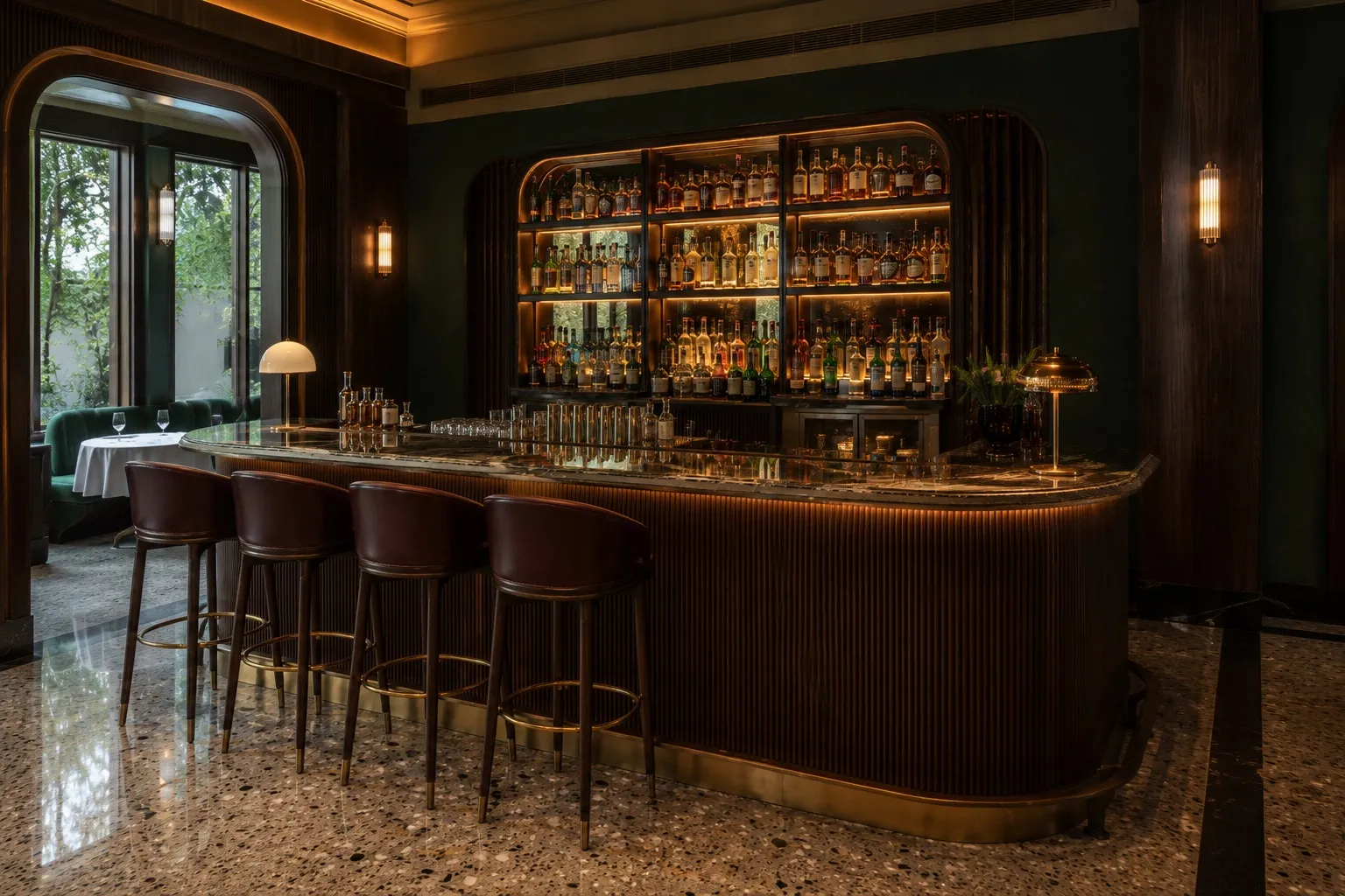 Curved fluted-wood bar with a brass foot rail and marble top, brown leather bar stools, a backlit shelf of bottles, and brass dome table lamps against a deep-green wall on a terrazzo floor