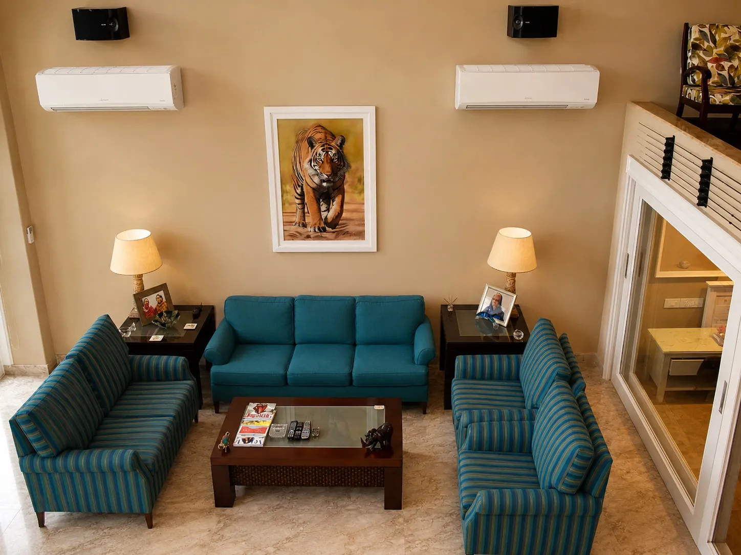 Family living room with a teal three-seater and striped sofas around a wood-and-glass coffee table, a framed tiger painting, and beige marble floor