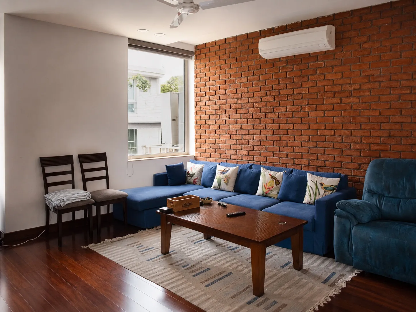 Family lounge with an exposed red-brick wall, a teal-blue fabric sectional and recliner, a wooden coffee table, and a striped dhurrie on dark hardwood