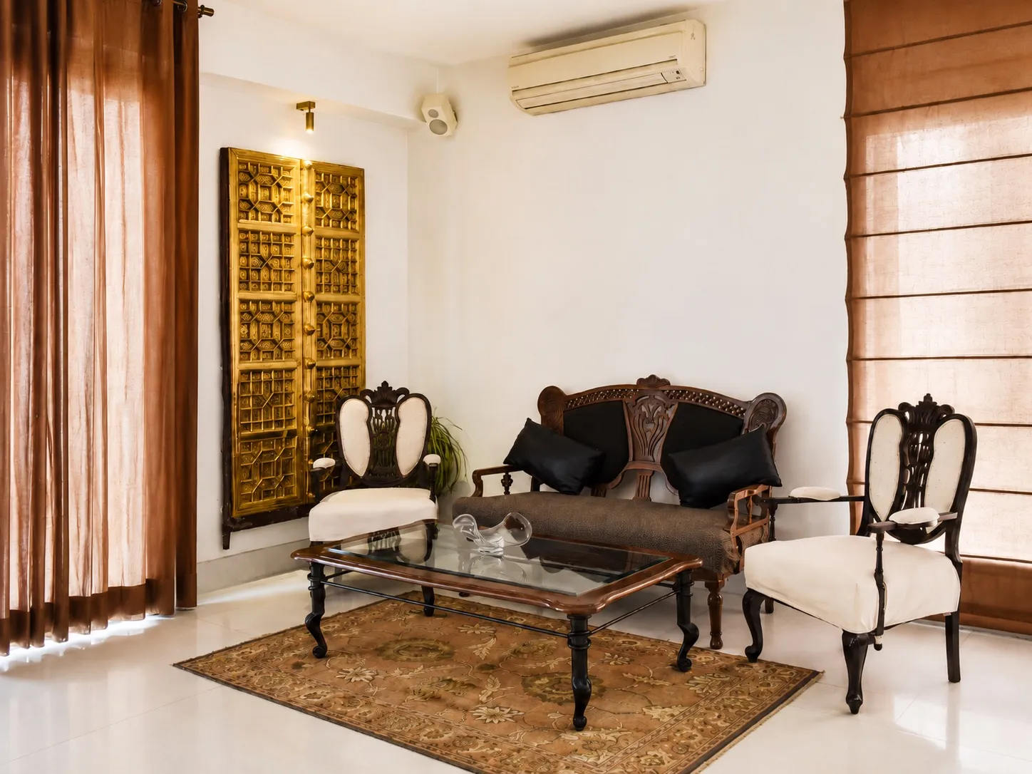 Formal sitting corner with an antique carved wooden settee, cream armchairs with black bolster cushions, and a large brass repoussé door panel mounted as wall art