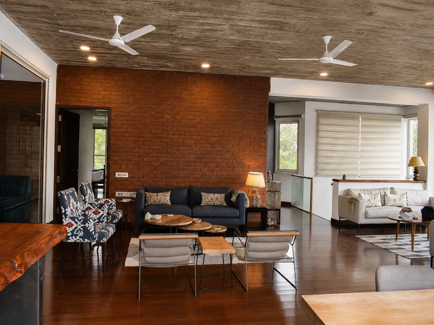 Open living area with a board-formed concrete ceiling and exposed red-brick feature wall, a navy sofa, ikat-print armchairs, round wooden nesting tables, and sling chairs