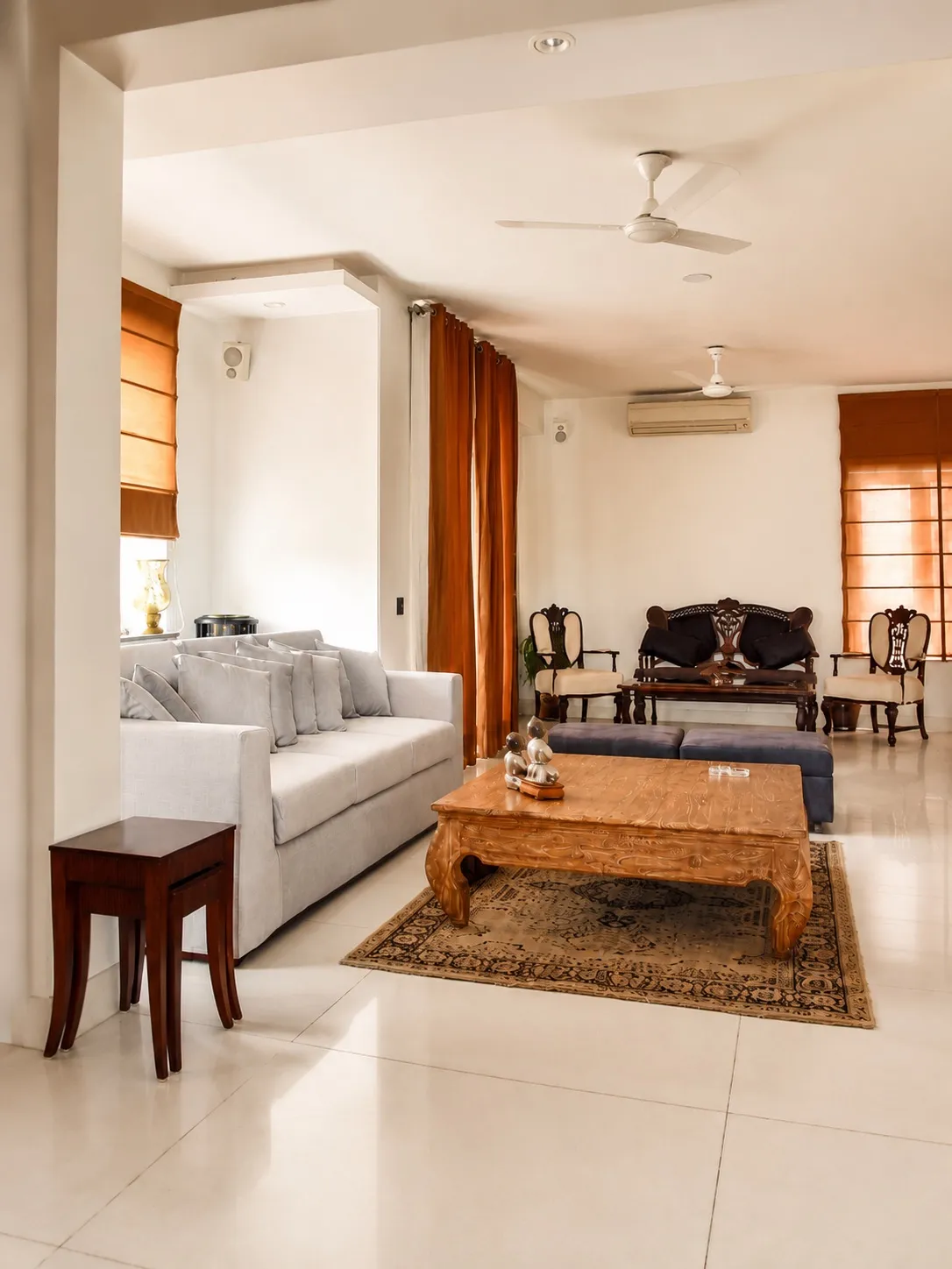 Bright lounge with a grey sofa, a carved gold-wood low table on a patterned rug, rust drapes, and antique carved seating on a white marble floor