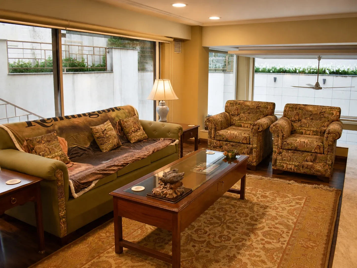 Media lounge with an olive-green sofa and safari animal-print armchairs, a wood-and-glass coffee table, a floral rug, and garden views
