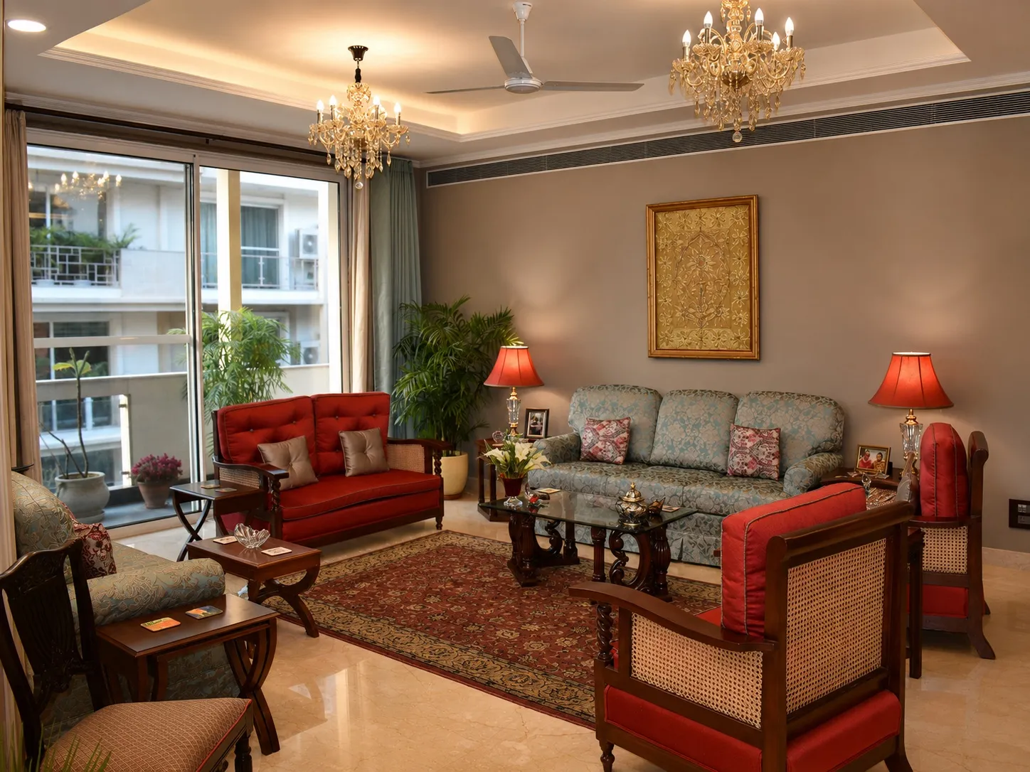 Formal living room with two crystal chandeliers, grey walls, red and floral-upholstered classical sofas, a gold framed artwork, and marble flooring opening to a balcony