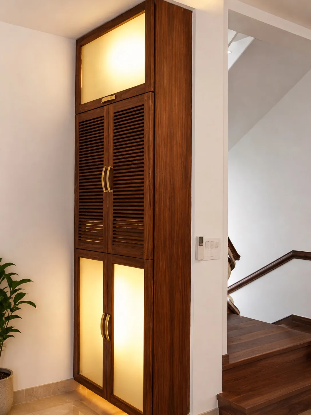 Floor-to-ceiling walnut storage unit with backlit frosted-glass and louvered panels beside a walnut staircase