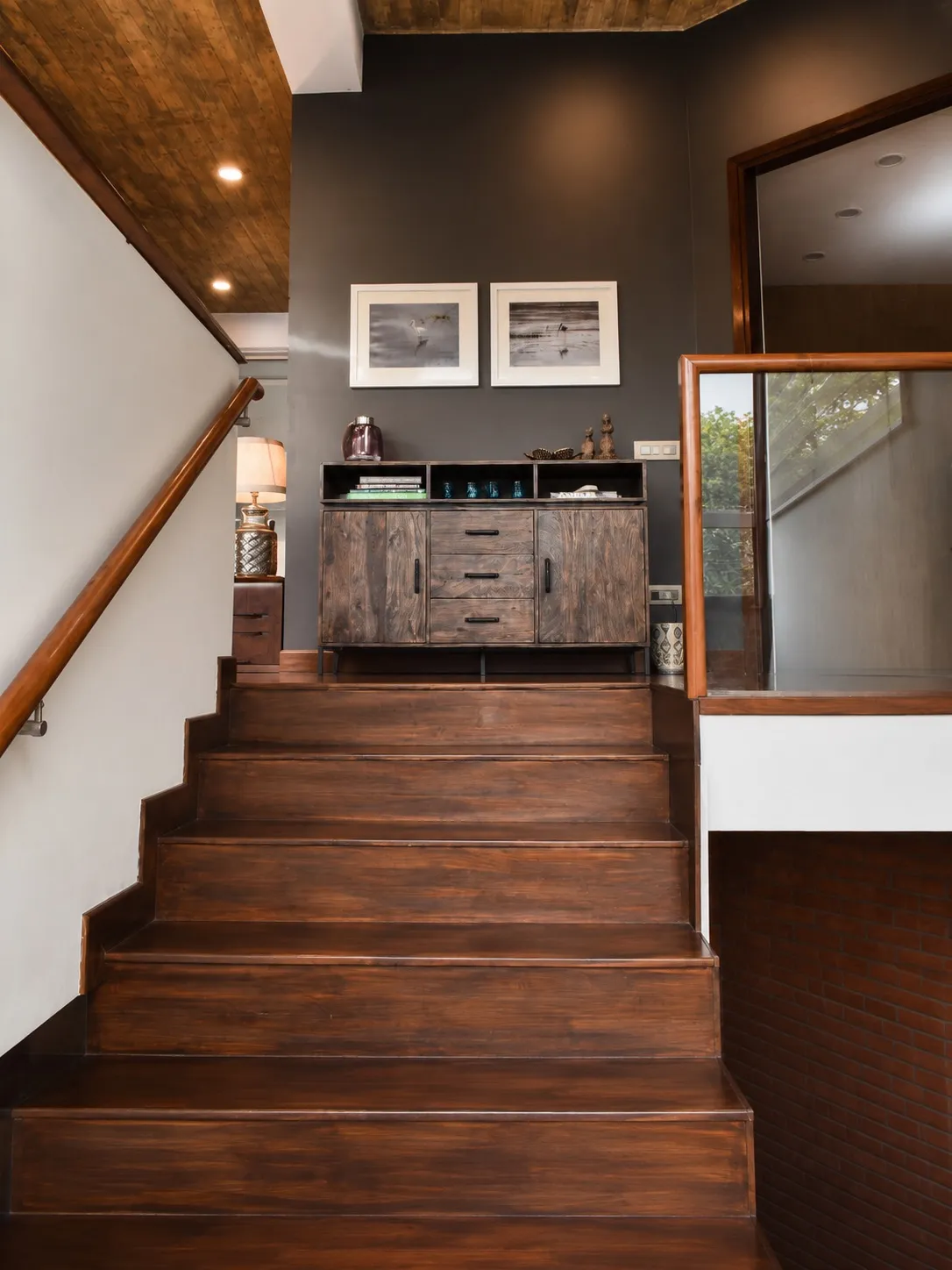 Timber staircase landing with a wood-slat ceiling, a reclaimed-wood sideboard and framed bird photographs on a charcoal wall, and a glass balustrade