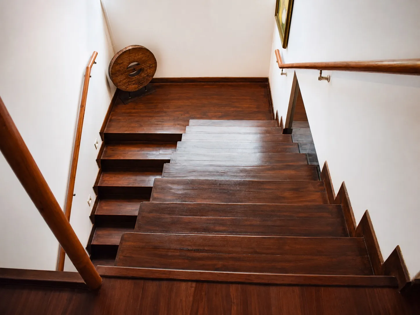 Dark-stained solid-wood staircase seen from above, with timber handrails on both sides and a carved wooden wheel artefact on the landing
