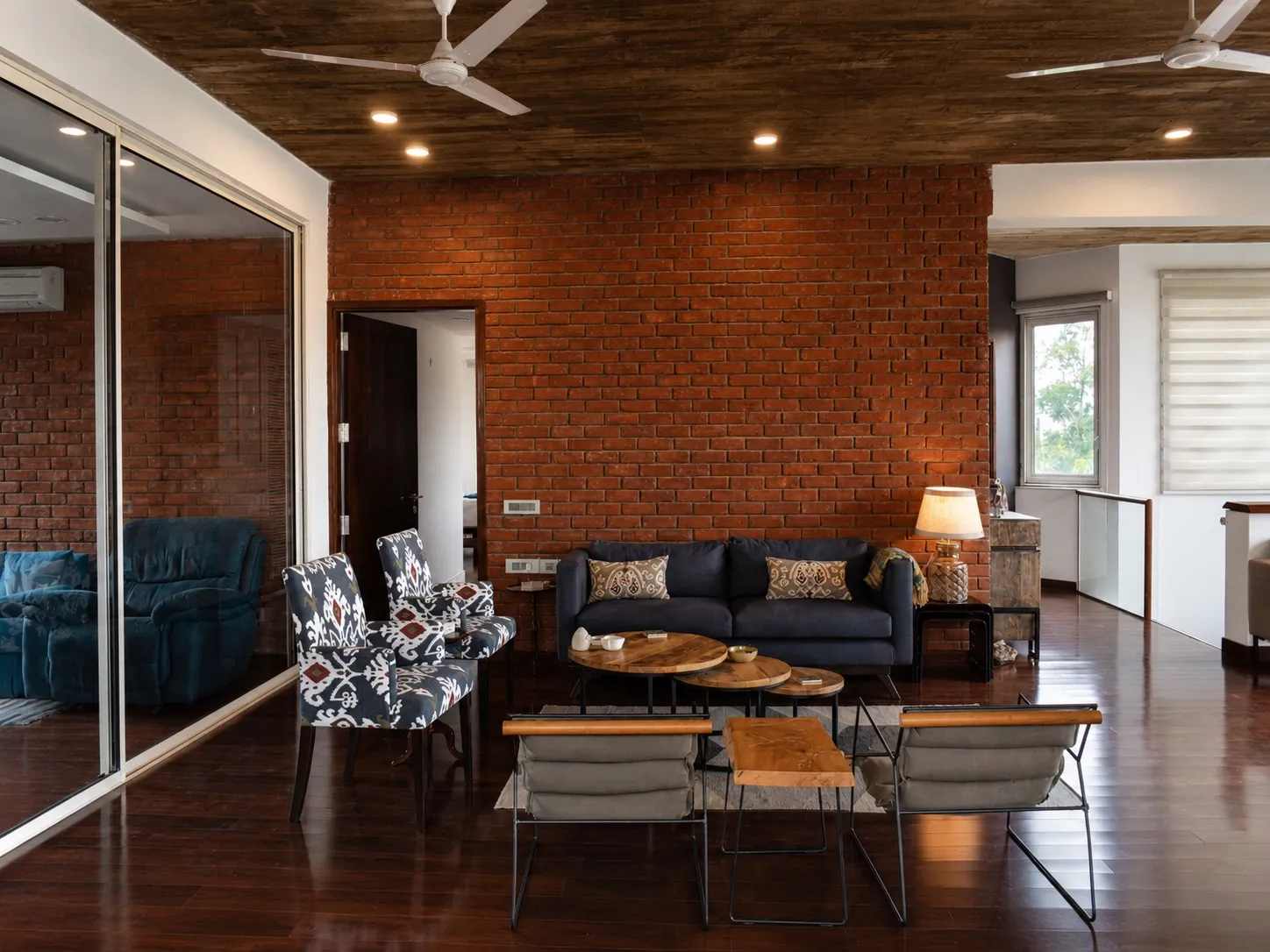 Living room with an exposed red-brick wall and concrete ceiling, a navy sofa with paisley cushions, ikat-print armchairs, round nesting tables, and a glazed partition to an adjoining lounge