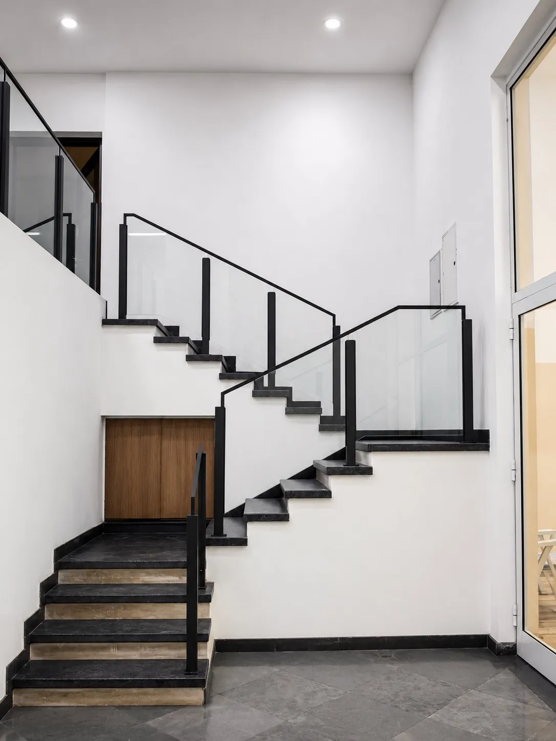 Double-height staircase with black stone treads and a black metal-and-glass balustrade against white walls, over a grey stone floor
