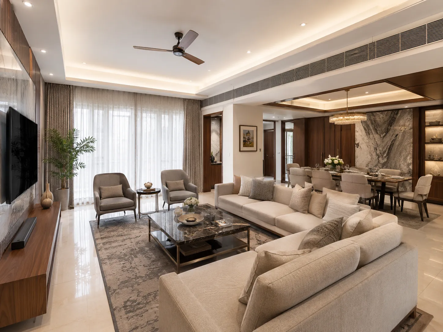 Open-plan living and dining room with beige marble floors, a cream sectional sofa, grey armchairs, a large marble TV backdrop, and a dining area with a crystal chandelier.