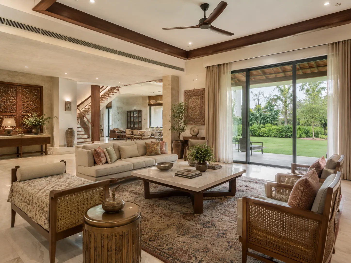 A spacious luxury living room with a square marble coffee table, beige sofas, cane-backed armchairs, a patterned rug, and large glass doors opening to a green lawn.