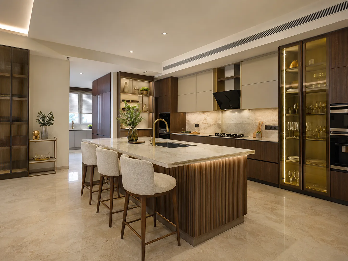 Wide view of a luxury layered kitchen featuring a central island with fluted wood base, a glass display cabinet with warm lighting, and a secondary wet kitchen in the background.