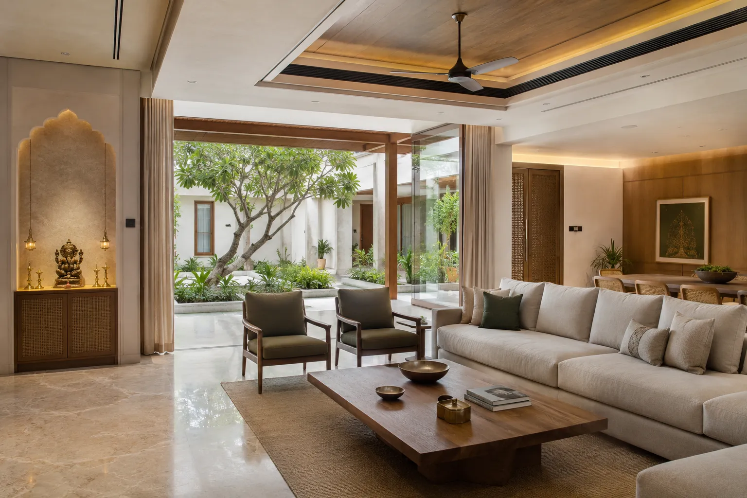 Large living room with marble floors, a solid wood coffee table, and neutral sofas facing an open central courtyard with a tree. A lit pooja niche with a Ganesha idol sits on the left.