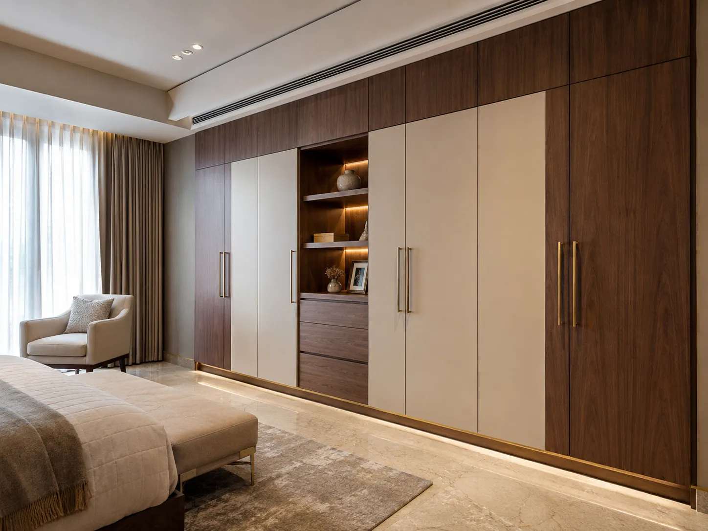 A luxurious bedroom featuring a large built-in wardrobe with dark wood framing, matte beige doors, brass handles, and a recessed toe kick illuminated by warm LED strip lighting on a polished marble floor.
