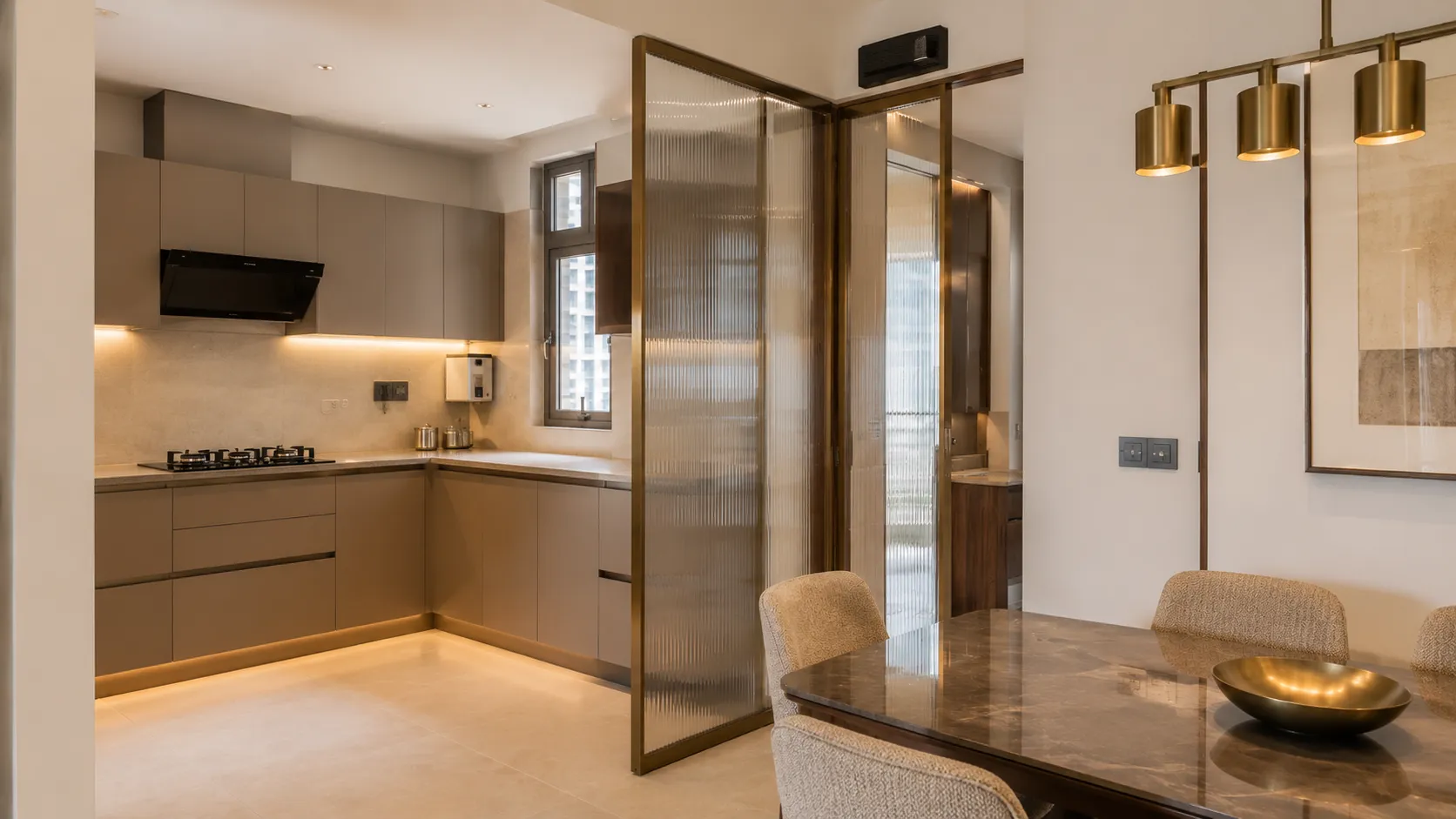Matte taupe modular kitchen with under-cabinet lighting, separated from a dining area by a brass-framed fluted glass partition.