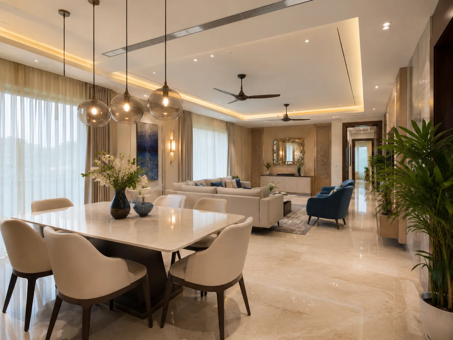 An open-plan dining and living area featuring a white marble dining table, beige chairs, glass globe pendants, and blue abstract wall art.