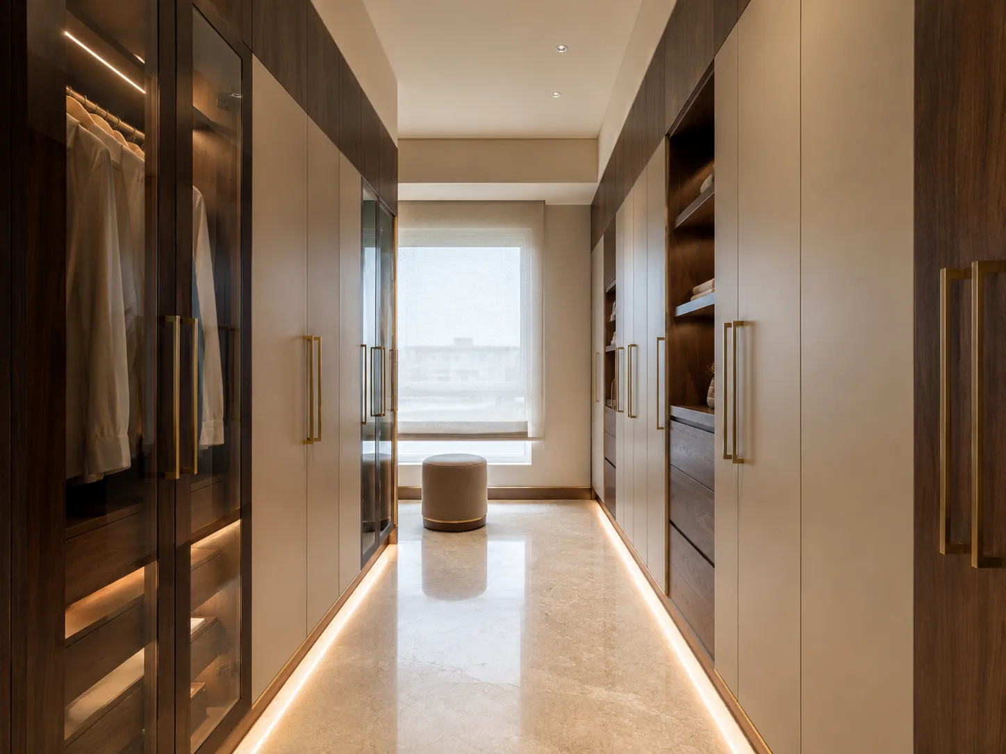 A walk-in closet corridor with sleek dark wood and beige cabinetry on both sides, featuring glass display doors, open shelving, and continuous under-cabinet toe kick lighting reflecting off the stone floor.