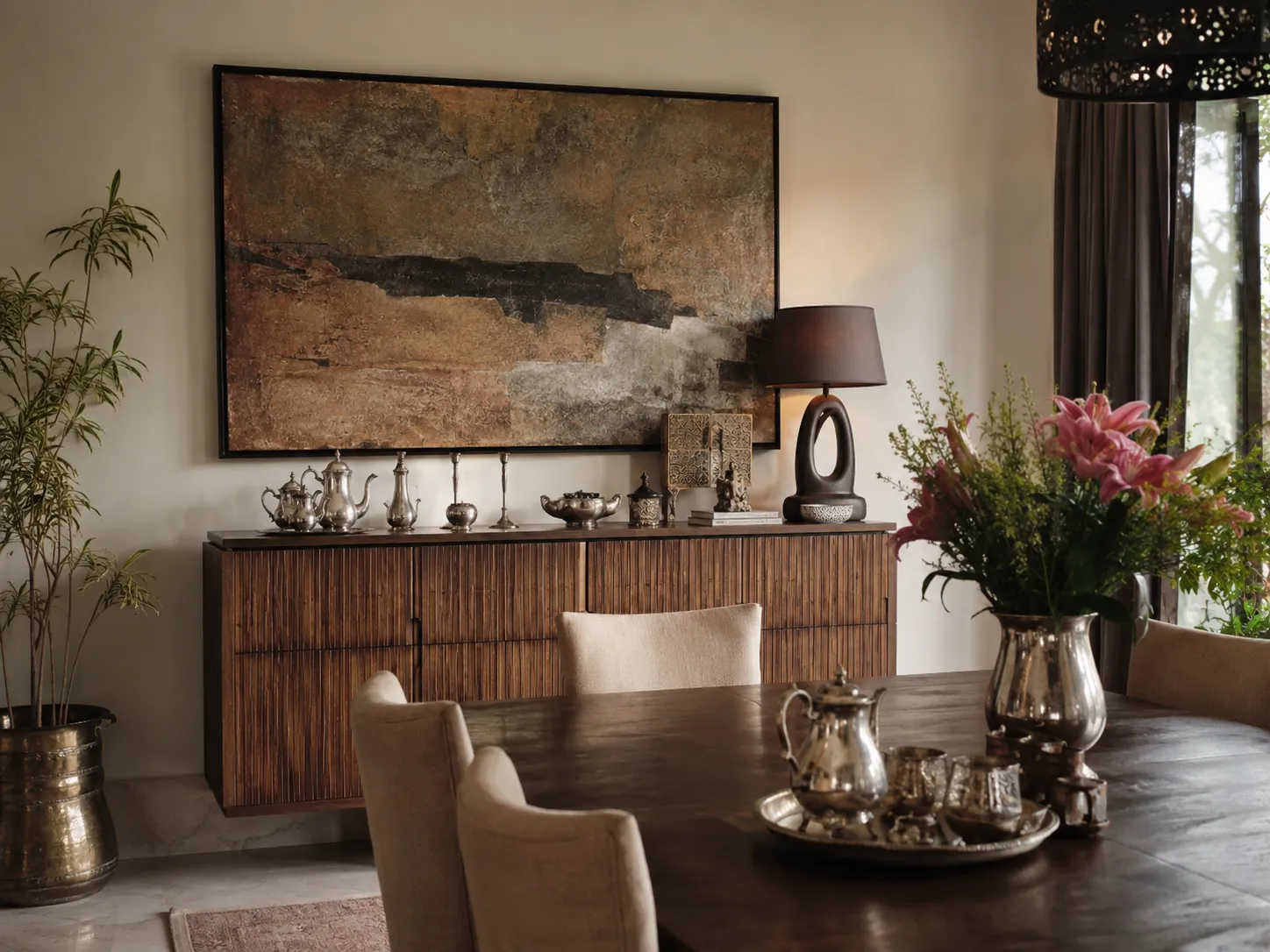 A dark wood dining table with a silver tea set, backed by a fluted wooden sideboard and a large textured earth-toned abstract painting.