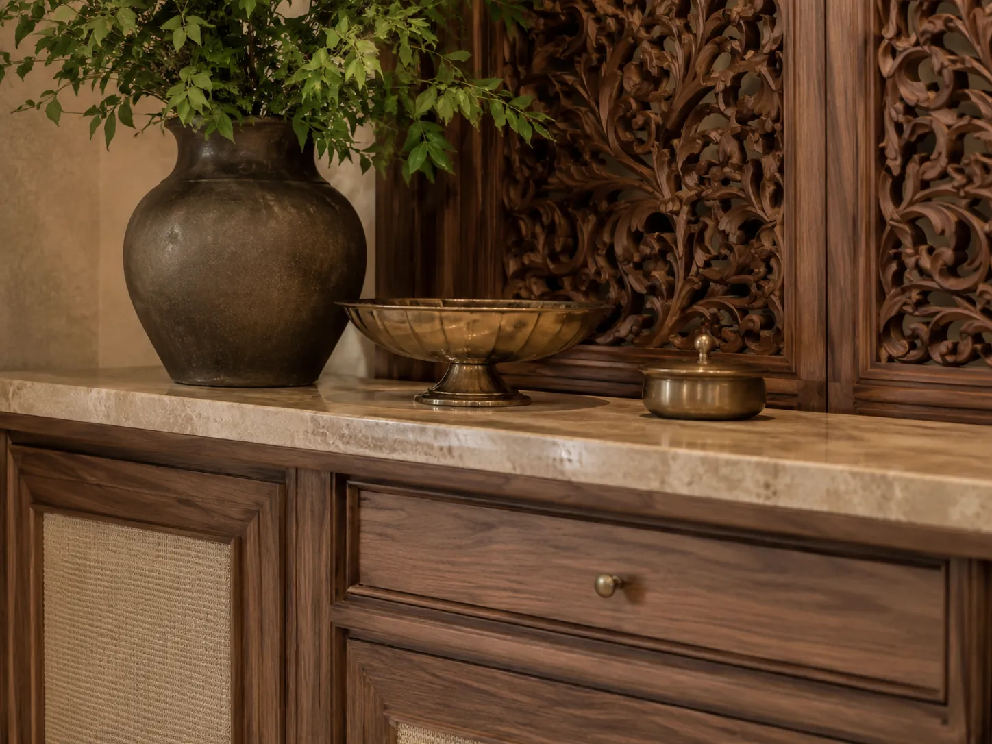 A close-up of a marble-topped wooden cabinet with cane inserts, set against an intricately carved wooden wall panel, styled with a dark vase and brass bowls.