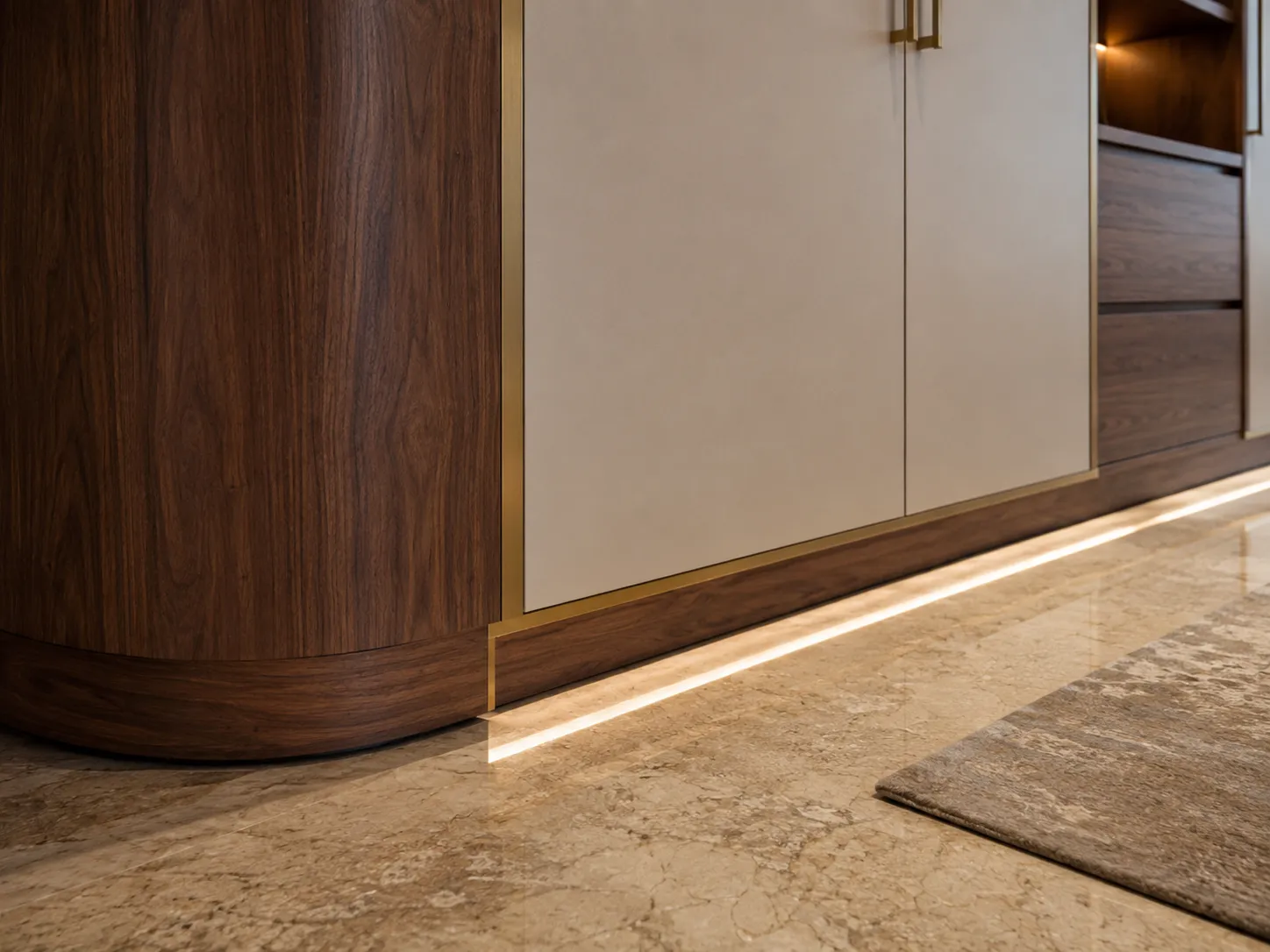 Close-up of a wardrobe base showing a curved dark wood end panel, matte beige doors with a thin brass trim, and a recessed wood toe kick with hidden LED lighting casting a glow on the marble floor.