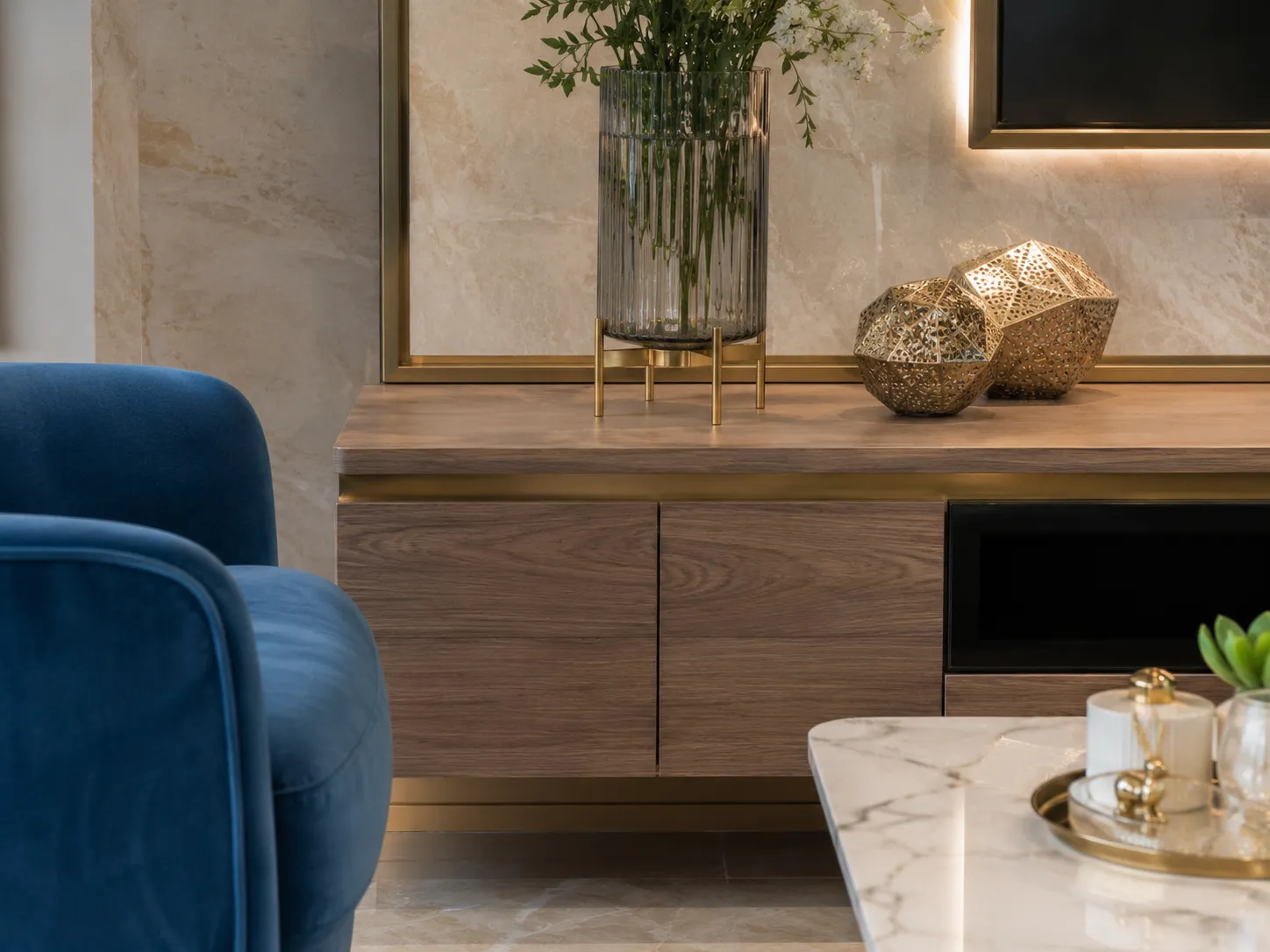 A close-up of a blue velvet armchair next to a wood and brass media console decorated with geometric gold accents and a fluted glass vase.