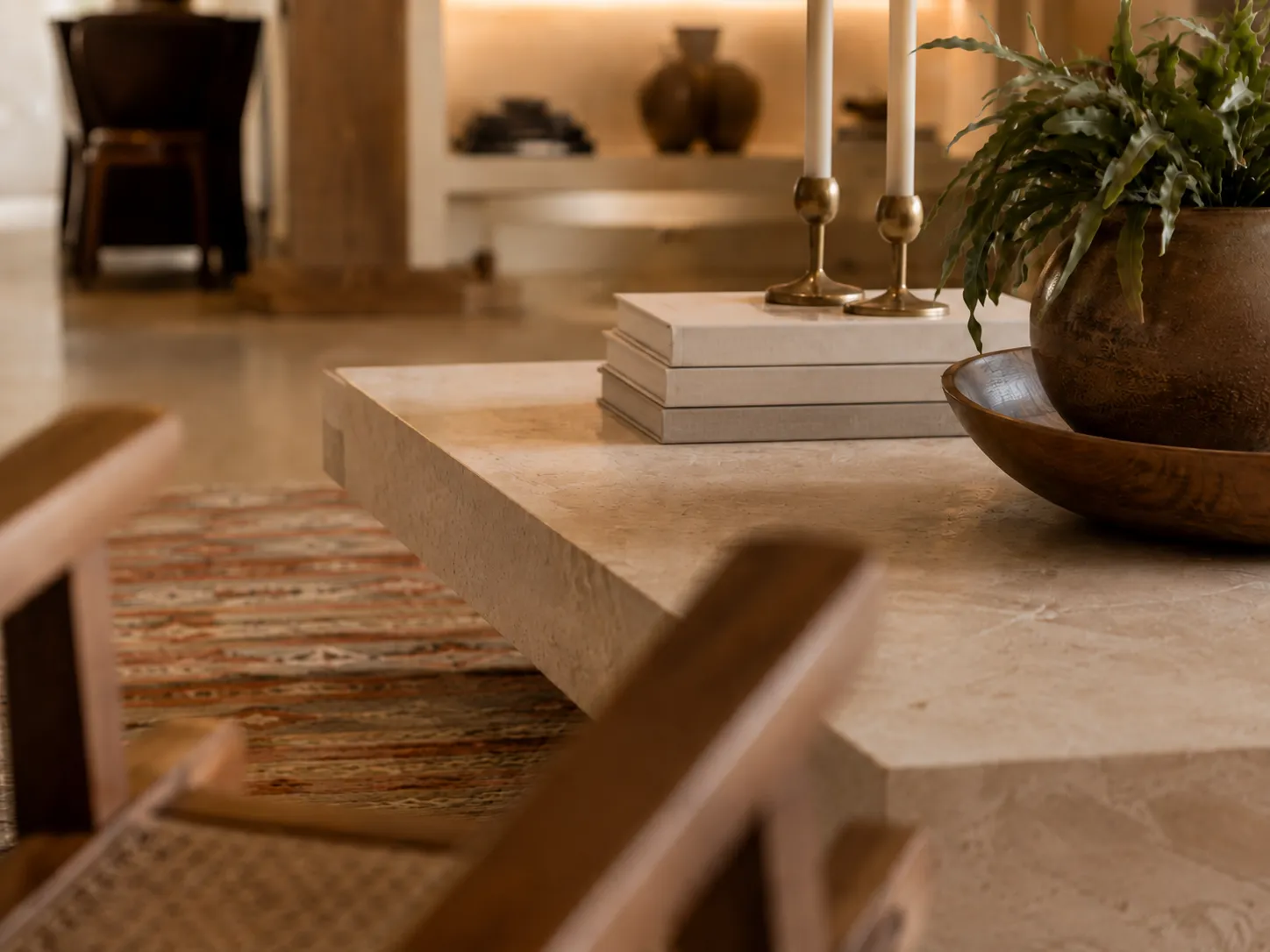 Close-up of a textured stone coffee table displaying stacked linen-bound books, two brass candlesticks with white taper candles, and a wooden bowl holding a potted green fern.
