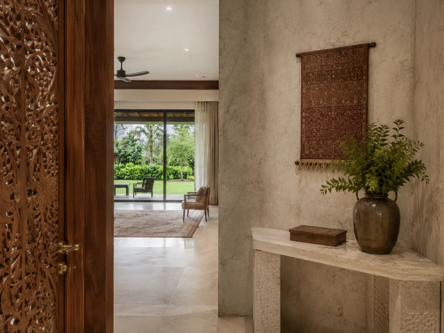 An entryway showing a heavily carved wooden door partially open to a bright living space, with a textured wall featuring a hanging tapestry and a stone console table.