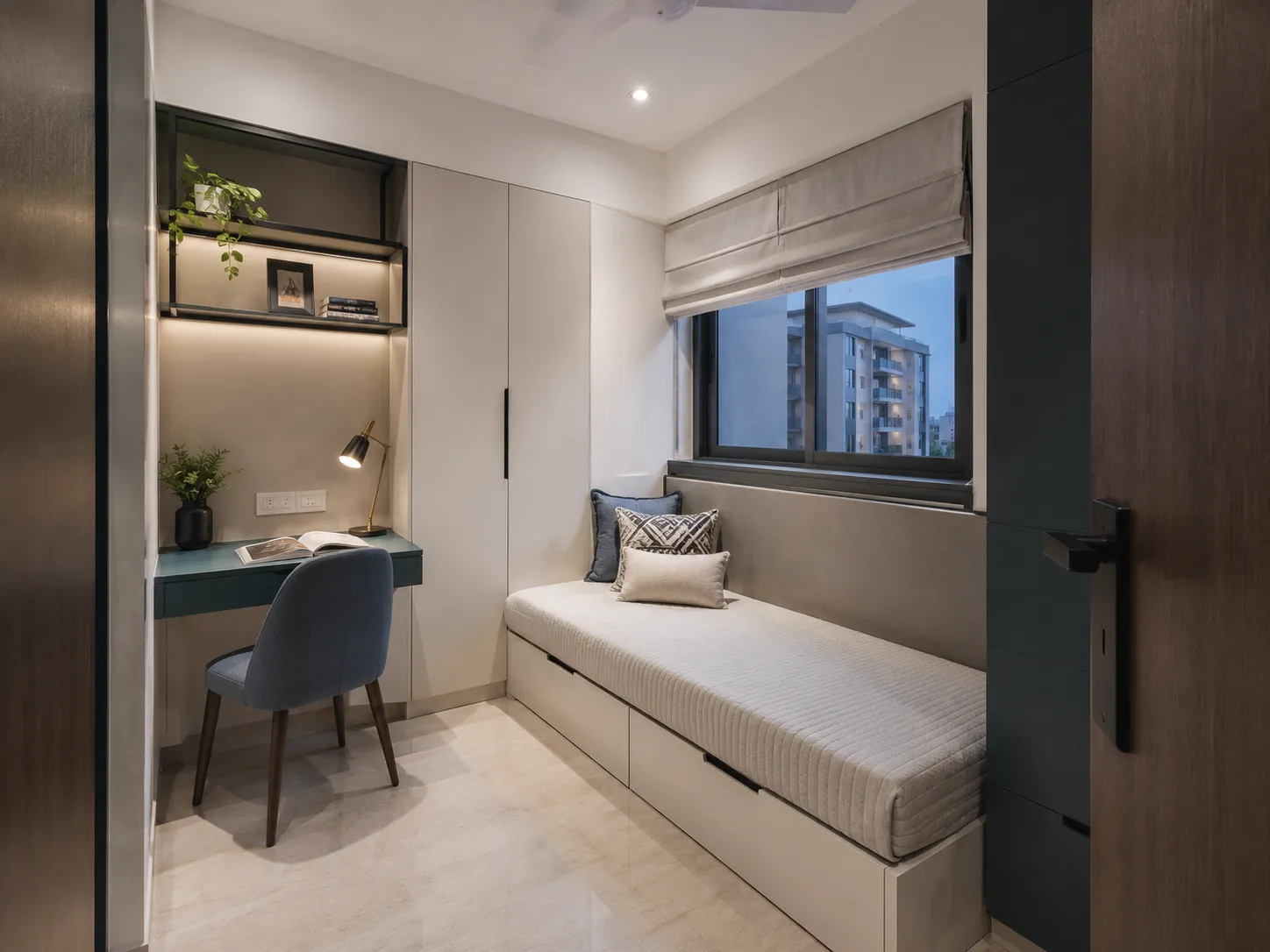 A compact room with a built-in daybed over storage drawers, a teal study desk, and open shelving illuminated by warm LED lights.