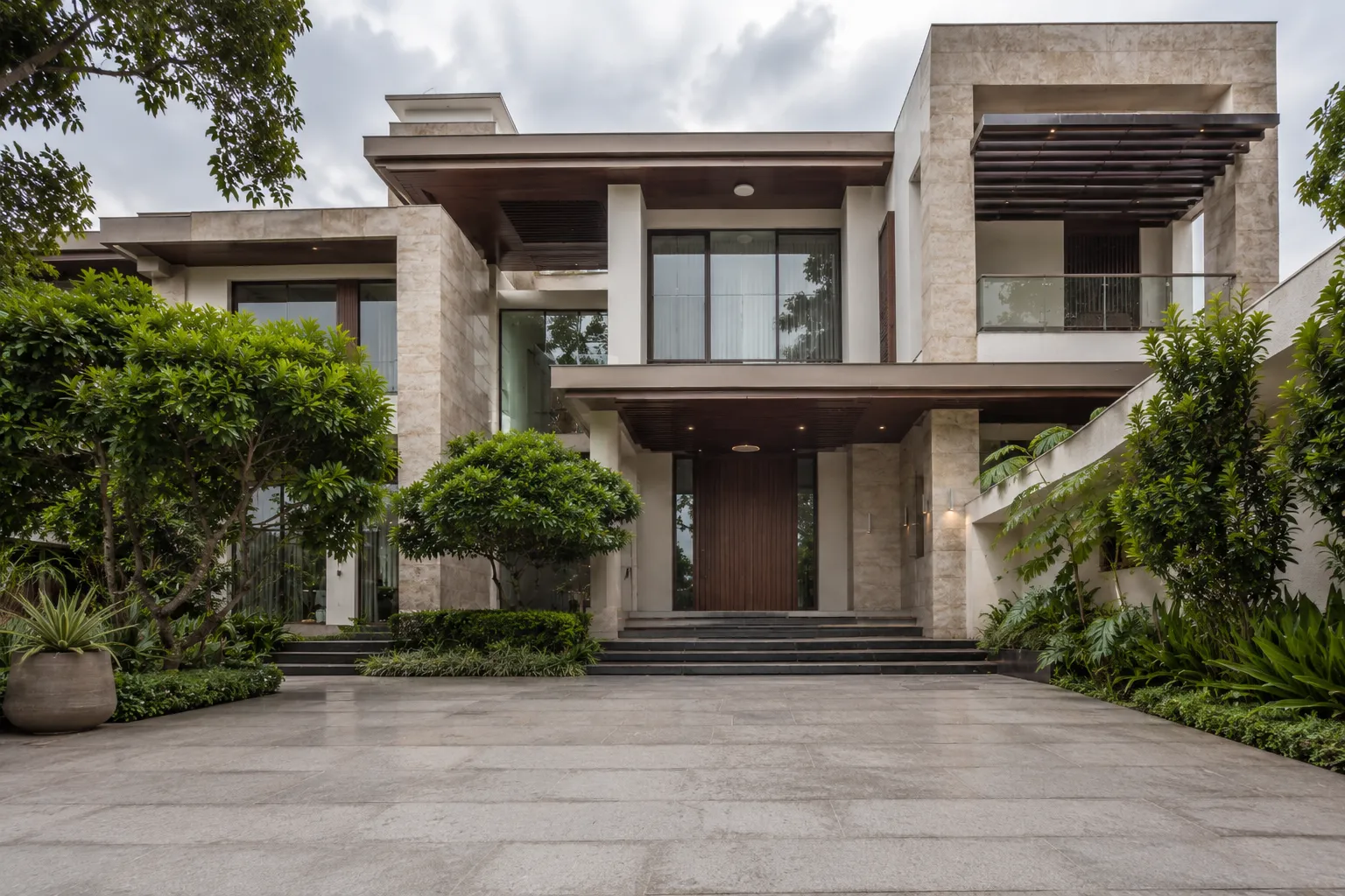 Exterior of a modern luxury villa showing stone cladding, dark wood overhangs, large glass windows, and a wide stone driveway.